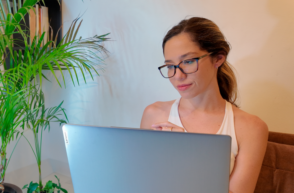 Fernanda Zely em seu computador, usando óculos e blusa branca, na foto compõe uma planta e uma estante de fundo.